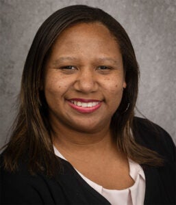 A woman with straight dark hair, wearing a black blazer and light blouse, smiles at the camera against a gray textured background.