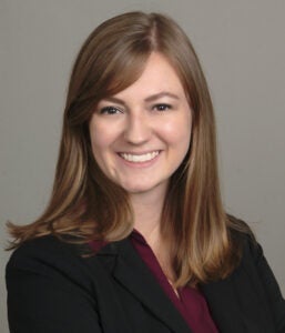 A woman with straight, light brown hair is smiling at the camera. She is wearing a dark blazer over a maroon top, posing in front of a plain, light background.