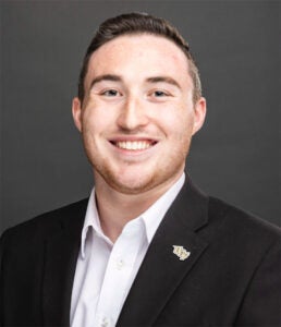 A young man with short brown hair in a black blazer and white shirt, smiling, posed against a plain dark background.