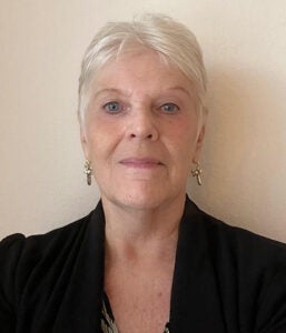 An older woman with short white hair, wearing earrings and a black top, poses in front of a plain light-colored wall.