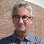A middle-aged man with gray hair and glasses is wearing a gray collared shirt and standing in front of a brick wall.
