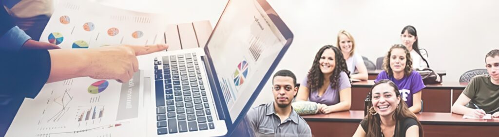 A person points at charts on paper next to a laptop displaying graphs; in the background, students sit in a classroom, facing forward and listening.