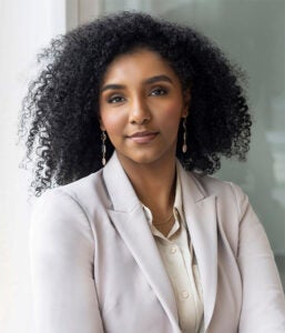 A woman with curly hair wearing a light-colored blazer and earrings stands indoors, looking confidently at the camera.