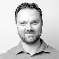 A man with short hair and a beard, wearing a collared shirt, faces the camera and smiles slightly against a plain background.