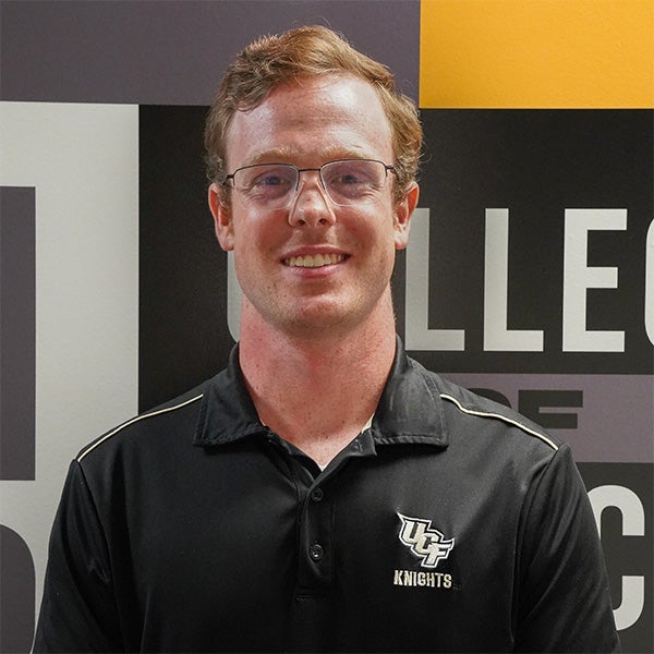 A man wearing glasses and a black UCF Knights polo shirt stands in front of a wall with partially visible text.