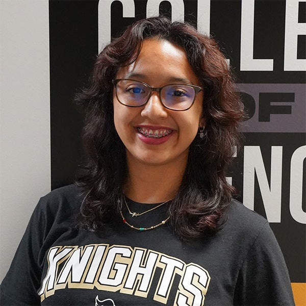 A person with wavy dark hair, glasses, and braces smiles while wearing a black "Knights" t-shirt, standing in front of a sign that partially reads "College of Engineering.