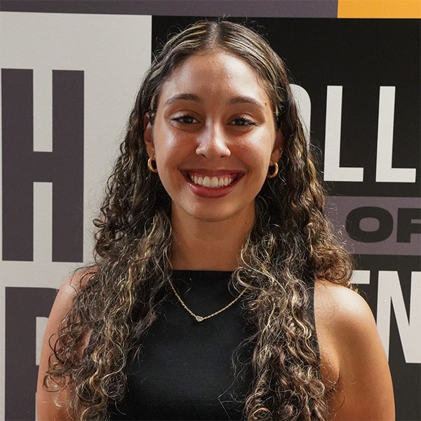 A woman with long, curly hair and a necklace stands smiling in front of a black and white background with text.