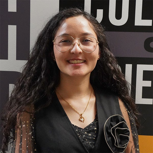 A young woman with long curly hair, glasses, and a black outfit with a decorative flower stands in front of a wall with bold text.