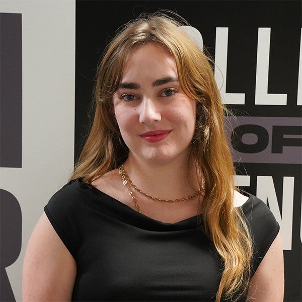 A person with long light brown hair wearing a black top and a gold necklace stands in front of a wall with bold black and white text.