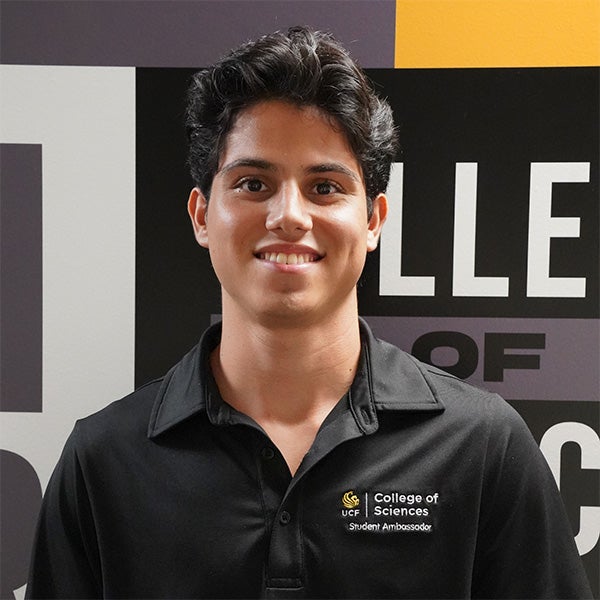 A young man wearing a black "College of Sciences Student Ambassador" polo shirt stands in front of a wall with bold, partially visible text.