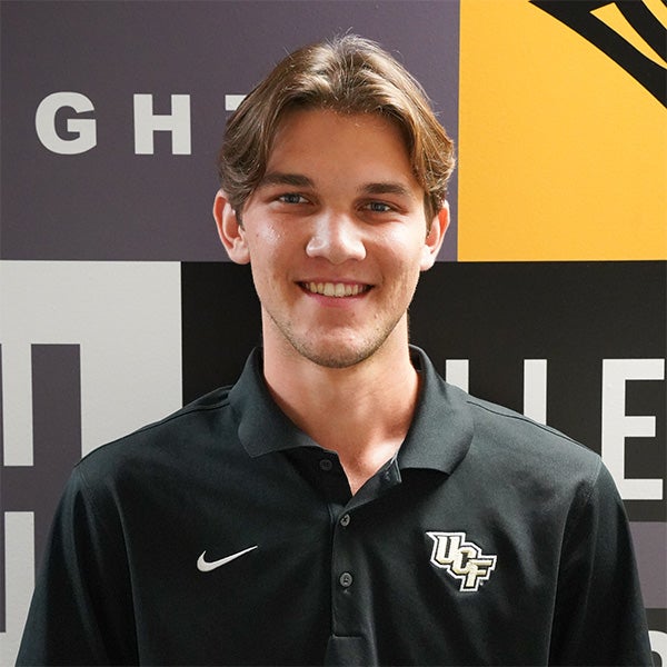 A young man with brown hair wearing a black collared shirt with the UCF logo stands in front of a wall with black, white, and yellow designs.