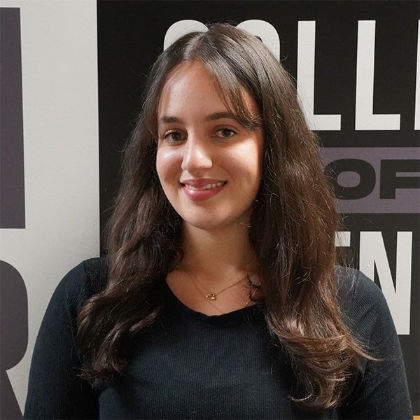 A woman with long brown hair wearing a black top stands in front of a wall with partially visible text. She is smiling at the camera.