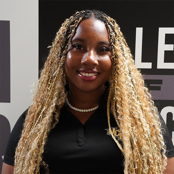 A woman with long, curly blonde hair wearing a black collared shirt with a UCF logo and a pearl necklace, standing in front of a sign with partial text.