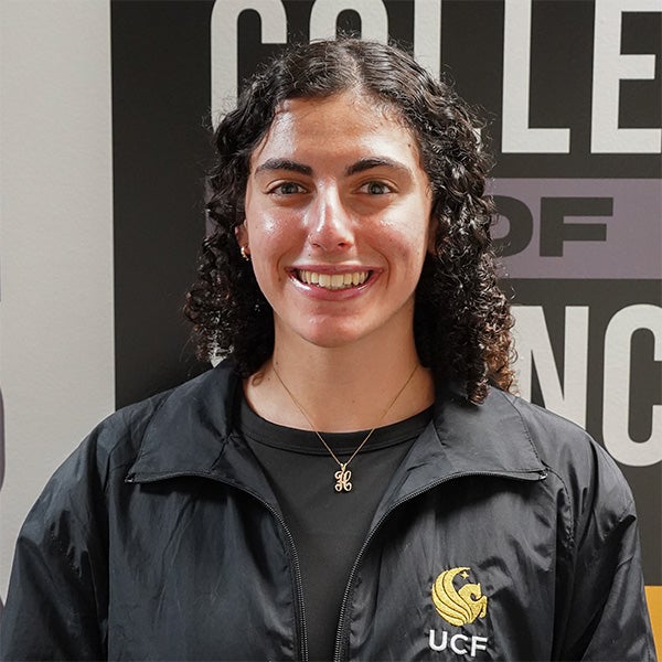 A person with curly dark hair smiles at the camera, wearing a black UCF jacket, standing in front of a sign that reads "College of Science.