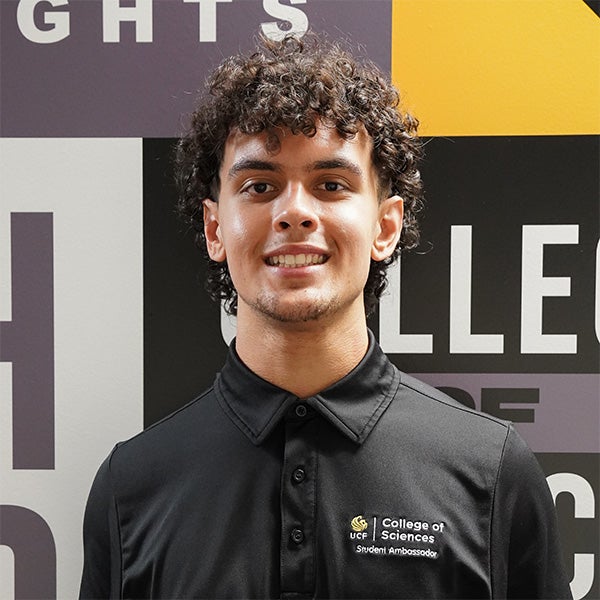 A person wearing a black "UCF College of Sciences Student Ambassador" polo shirt stands in front of a graphic wall with large text.