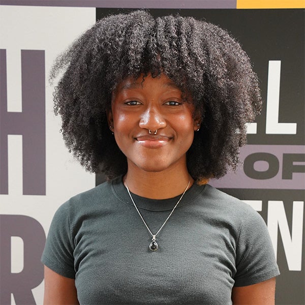 A young woman with natural curly hair, wearing a dark t-shirt and a pendant necklace, stands in front of a wall with large, bold text.