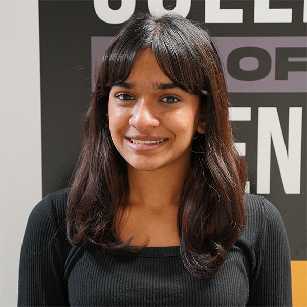 A young woman with long dark hair and a black top smiles in front of a sign with large white and colored block letters.