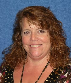 A woman with curly, shoulder-length brown hair smiles at the camera against a blue background.