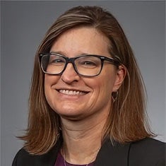 A woman with straight, shoulder-length brown hair and glasses smiles at the camera, wearing a dark blazer and a purple top, against a plain gray background.