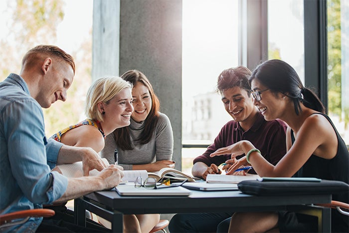 Five young adults sit at a table, smiling and working together with notebooks and papers in a brightly lit room.