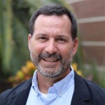 A man with short dark hair and a beard is smiling at the camera, wearing a light blue shirt and a dark jacket. The background is blurred with greenery and a brick wall.