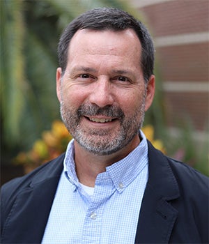 A man with short dark hair and a beard is smiling at the camera, wearing a light blue shirt and a dark jacket. The background is blurred with greenery and a brick wall.