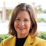 Woman with shoulder-length brown hair wearing a yellow blazer and black top, smiling at the camera outdoors with a blurred background.