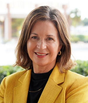 Woman with shoulder-length brown hair wearing a yellow blazer and black top, smiling at the camera outdoors with a blurred background.