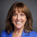 Woman with shoulder-length brown hair, wearing a blue blazer and pearl earrings, smiling at the camera with a gray background.