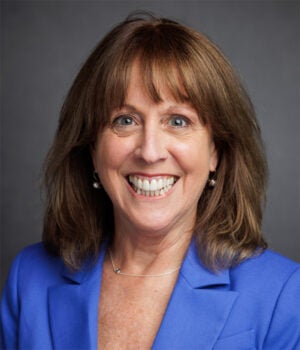 Woman with shoulder-length brown hair, wearing a blue blazer and pearl earrings, smiling at the camera with a gray background.