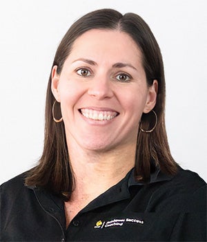 A woman with straight brown hair and hoop earrings smiles at the camera, wearing a black collared shirt against a plain light background.