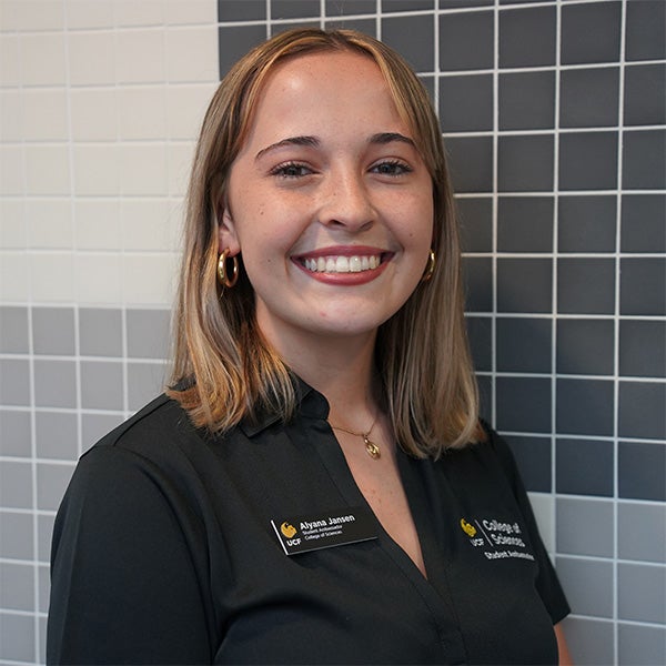 A person with shoulder-length blond hair smiles at the camera, wearing a black collared shirt and name tag, standing in front of a wall with black, gray, and white tiles.