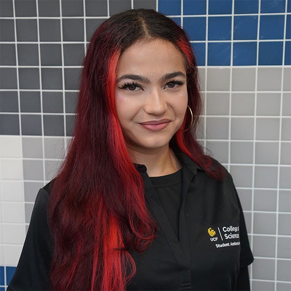 Woman with long red and black hair wearing a black UCF College of Sciences shirt stands in front of a tiled wall with shades of blue, gray, and white.