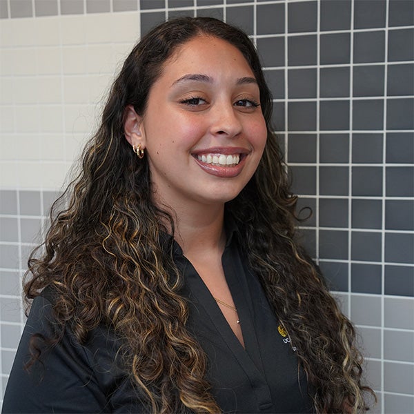 A woman with long curly hair smiles while standing in front of a tiled wall with white, gray, and black squares. She is wearing a black collared shirt and small hoop earrings.