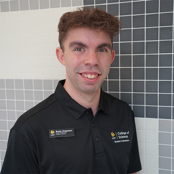 A man wearing a black “College of Sciences” polo and name badge stands in front of a black and white tiled wall, smiling at the camera.