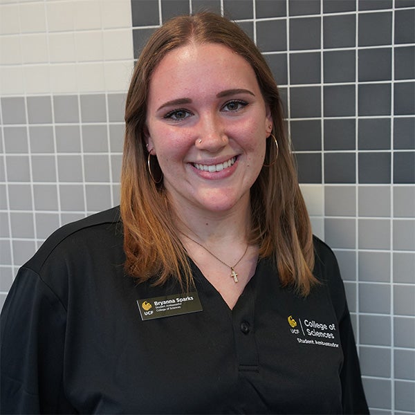 A woman with shoulder-length hair wearing a black UCF College of Sciences shirt and name tag stands in front of a gray and white tiled wall, smiling at the camera.