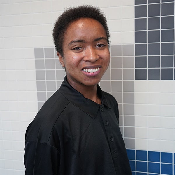Person with short hair wearing a black collared shirt stands in front of a wall with white, grey, and blue tiles, smiling at the camera.