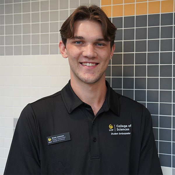 A young man with short brown hair smiles, wearing a black “College of Sciences Student Ambassador” polo and nametag, standing before a tiled wall with gray, white, and yellow squares.