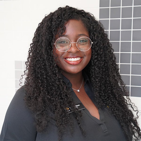 Woman with long curly hair and round glasses smiles at the camera, wearing a black shirt and name tag, standing in front of a tiled wall.
