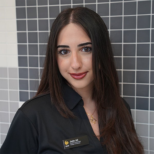 Woman with long dark hair wearing a black collared shirt and name tag stands in front of a black and white tiled wall, facing the camera and smiling slightly.