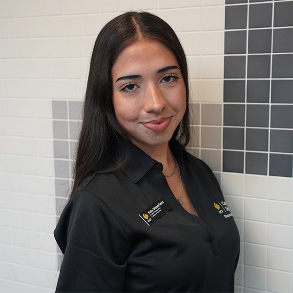 Woman with long dark hair wearing a black collared shirt and name tag stands in front of a tiled wall with black, gray, and white square patterns.