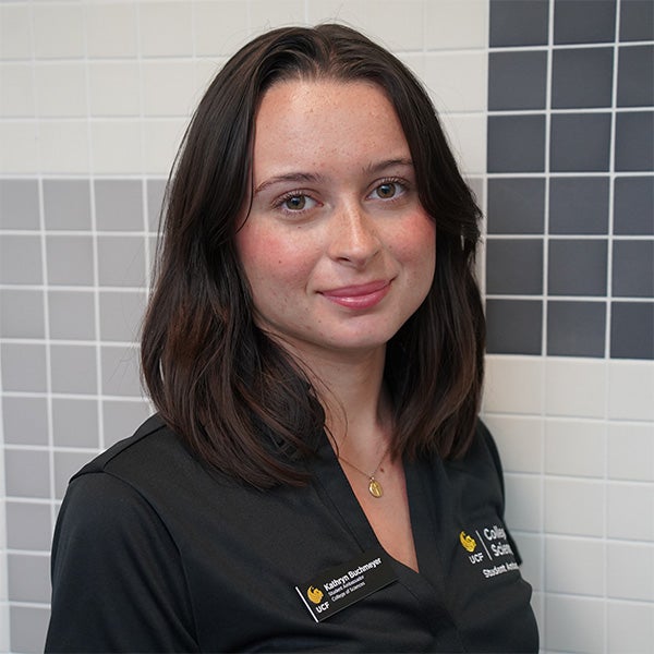 Woman with shoulder-length brown hair wearing a black collared shirt, standing in front of a tiled wall with a neutral expression.