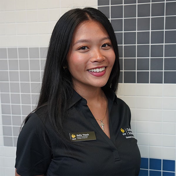 A woman with long black hair, wearing a black collared shirt and name badge, stands and smiles in front of a tiled wall with a grid pattern.