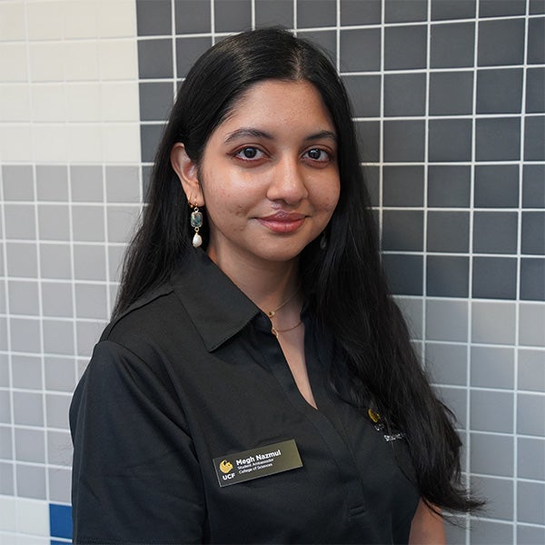Woman with long dark hair, wearing a black collared shirt and name tag, stands in front of a tiled wall with various shades of gray and white tiles.