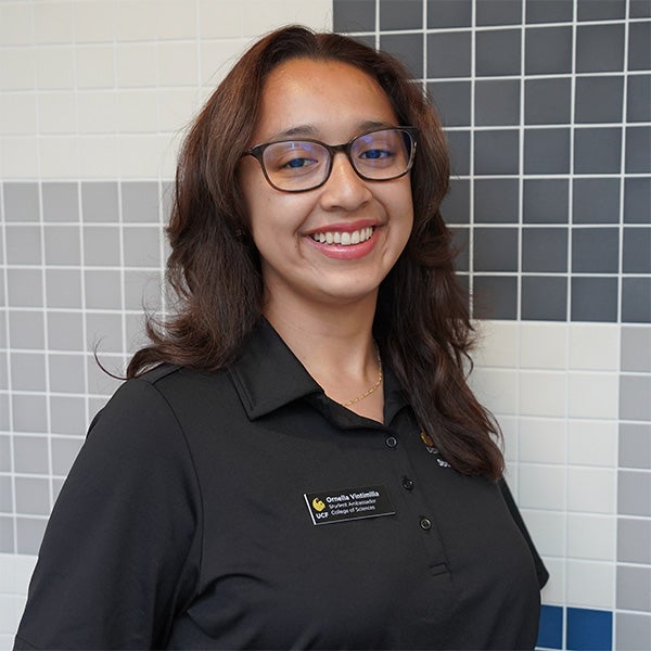 Person wearing glasses and a black collared shirt with a name badge stands in front of a tiled wall, smiling at the camera.