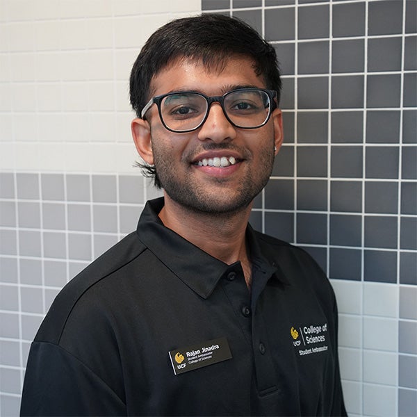 A person wearing glasses and a black “College of Sciences” polo shirt with a name tag stands in front of a tiled wall, smiling at the camera.