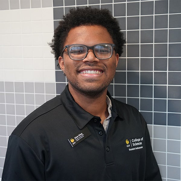 A person wearing glasses and a black College of Sciences shirt smiles while standing in front of a gray and white tiled wall.