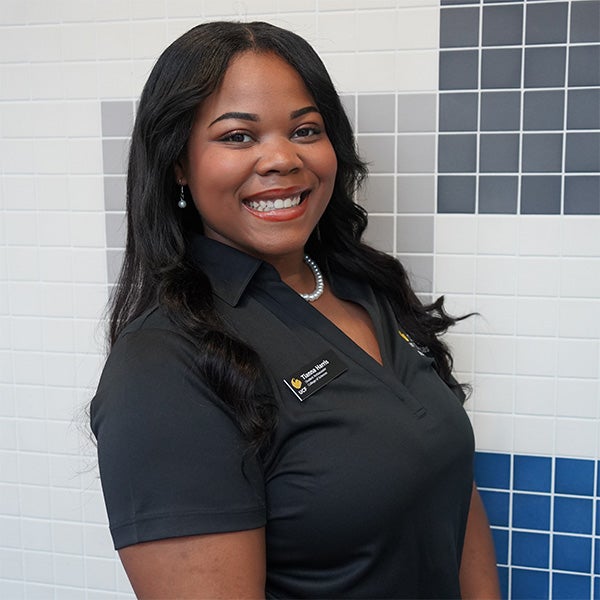 A woman with long dark hair smiles at the camera, standing in front of a tiled wall. She wears a black collared shirt and a name badge.