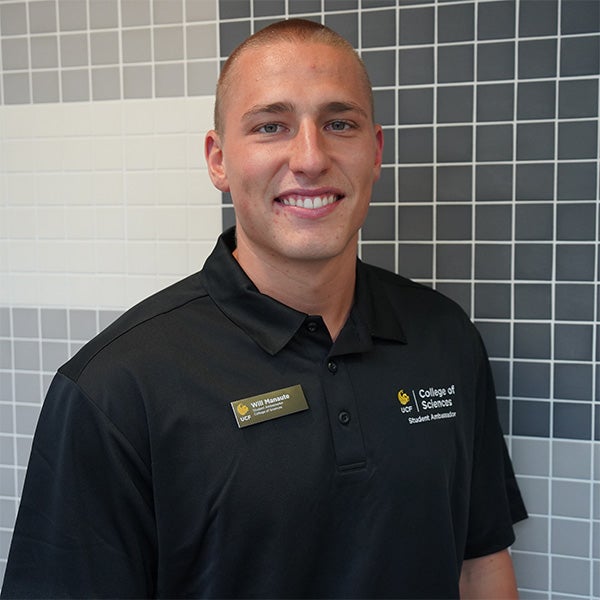 A man in a black polo shirt with a name tag stands in front of a tiled wall, smiling at the camera.